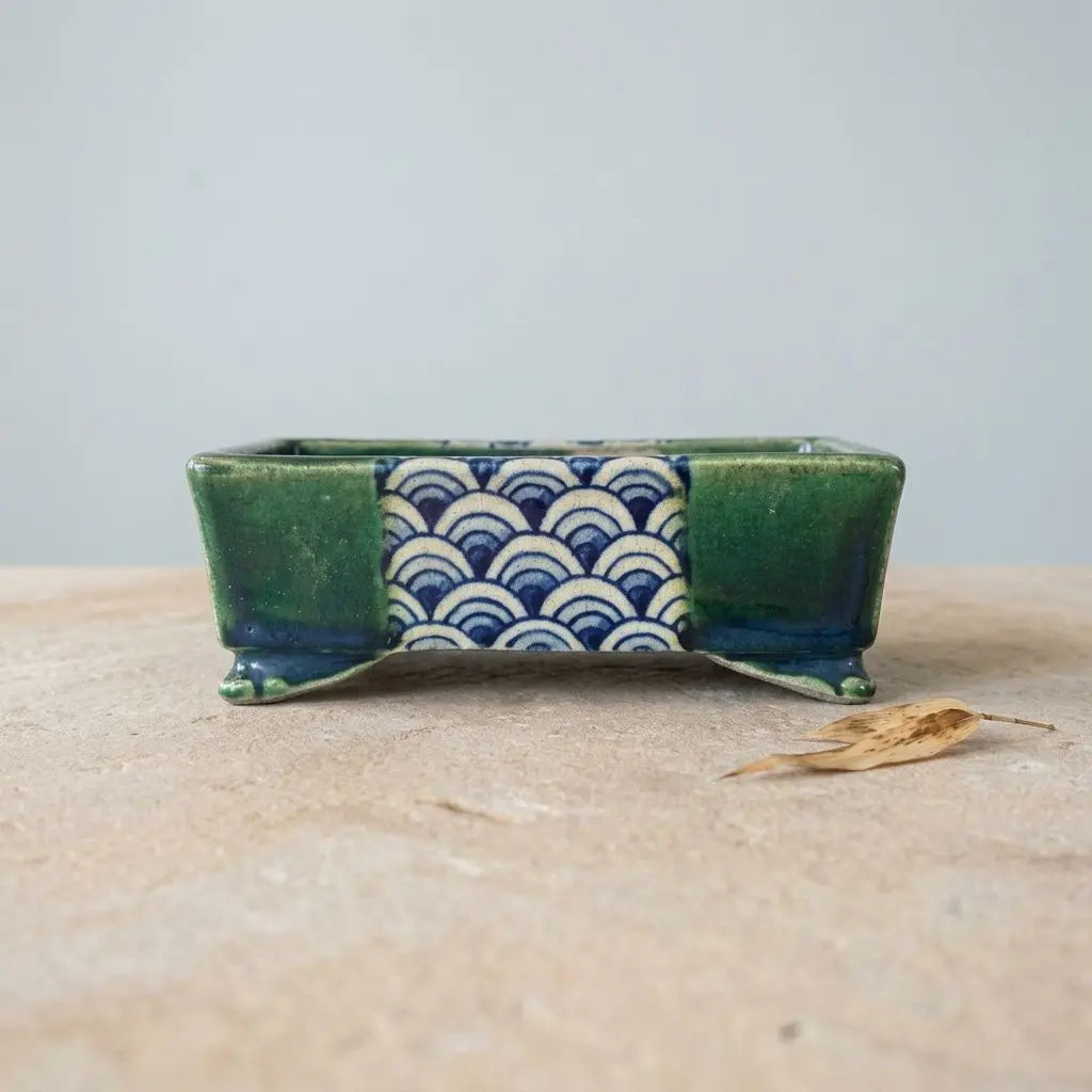 Petite soucoupe en poterie, motif géométrique en vague bleu sur fond vert, posé sur une surface beige avec une feuille sèche à côté. Pot bonsaï Terahata Satomi.