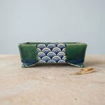 Petite soucoupe en poterie, motif géométrique en vague bleu sur fond vert, posé sur une surface beige avec une feuille sèche à côté. Pot bonsaï Terahata Satomi.